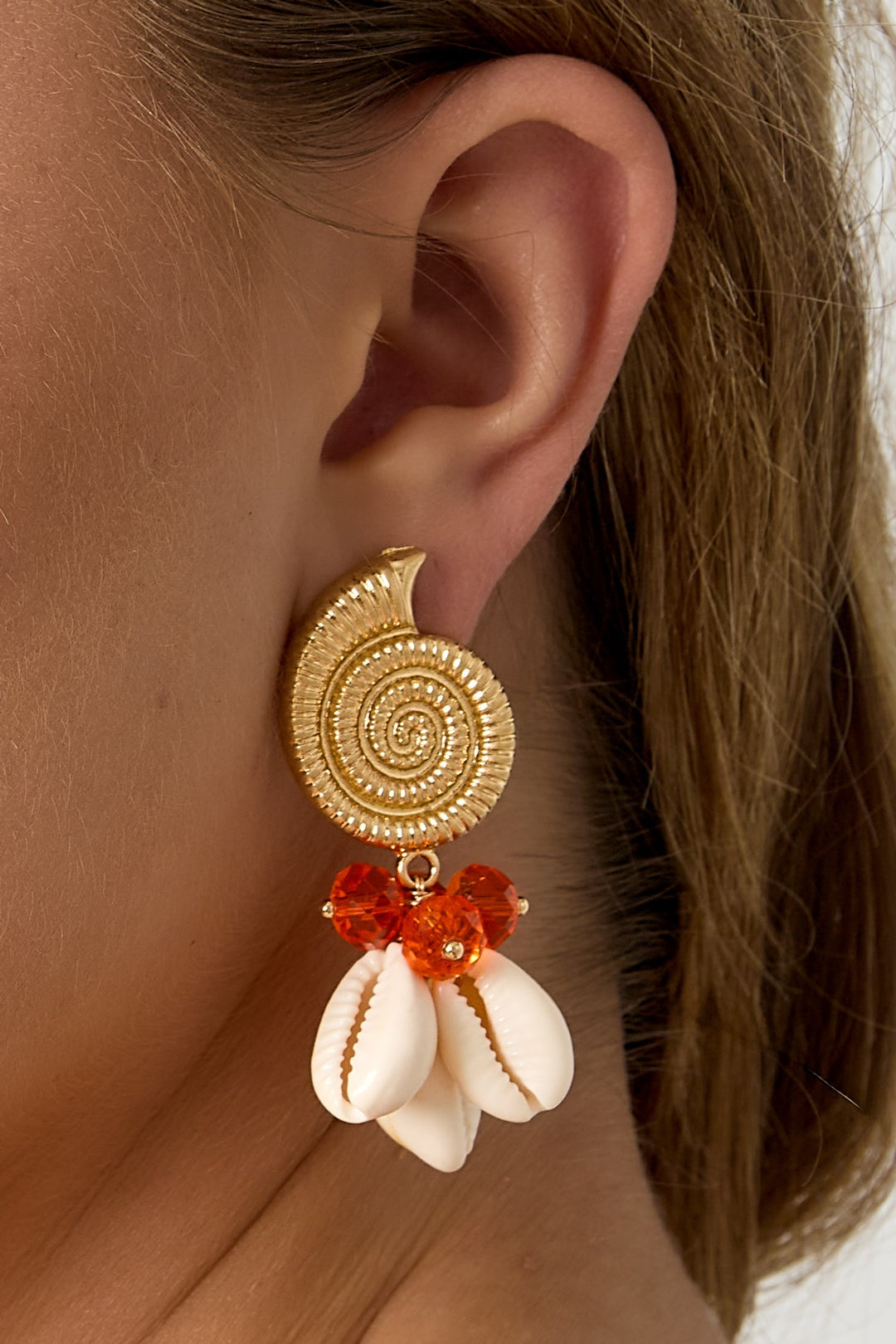 Close-up side view of woman wearing gold spiral shell earrings with orange glass beads and cowry shells
