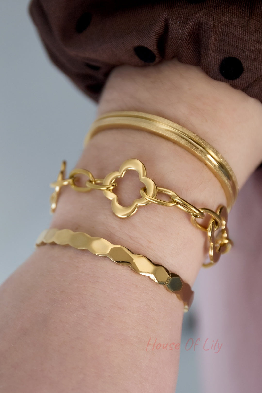 Wrist wearing a layered bracelet set including a gold scalloped edge bangle, a floral-link chain bracelet, and a smooth gold bangle, styled with a brown polka-dot sleeve