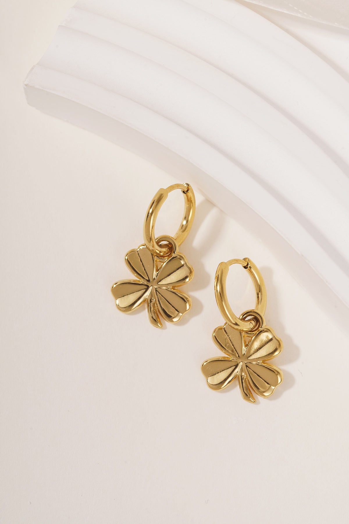 Close-up of gold four-leaf clover hoop earrings resting on a white textured surface