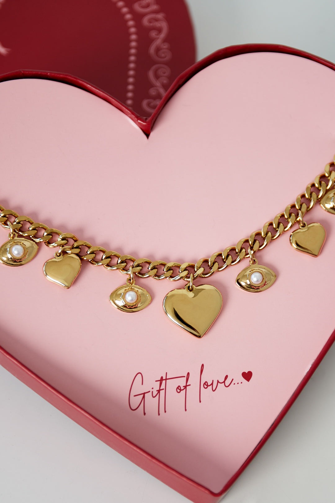 Close-up of gold-tone charm bracelet showing heart and pearl-adorned eye charms on a chunky chain inside pink heart box.