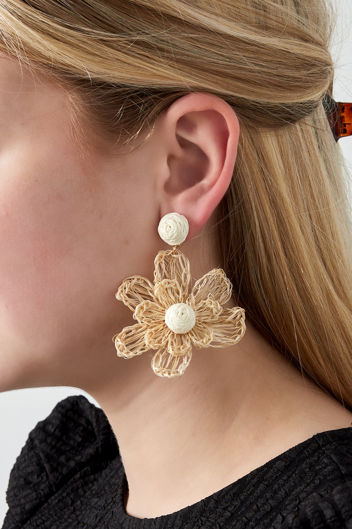 Close-up side view of a woman wearing a cream-colored raffia floral dangle earring with round woven stud and natural petal details