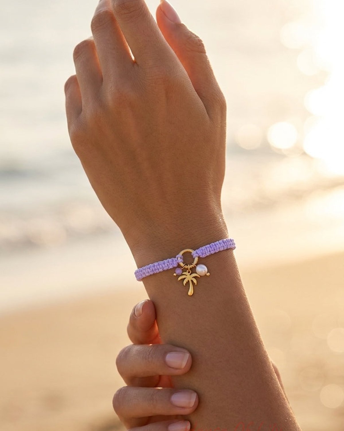 A light purple woven macrame bracelet with a gold palm tree charm and pearl on a beach.