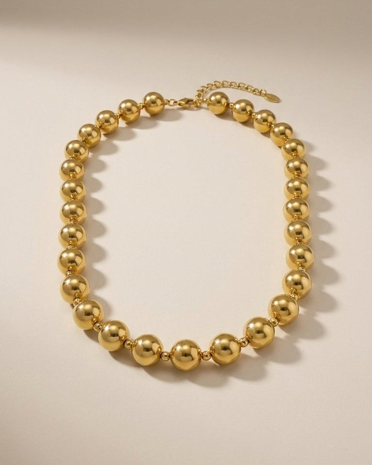 A gold beaded necklace laid flat in a circle on a neutral surface, featuring reflective metallic spheres.