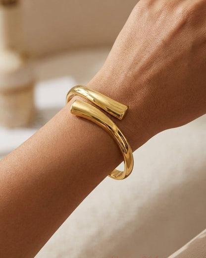 A polished gold cuff bracelet worn on a person's wrist, featuring a thick, minimalist wrap-around design.