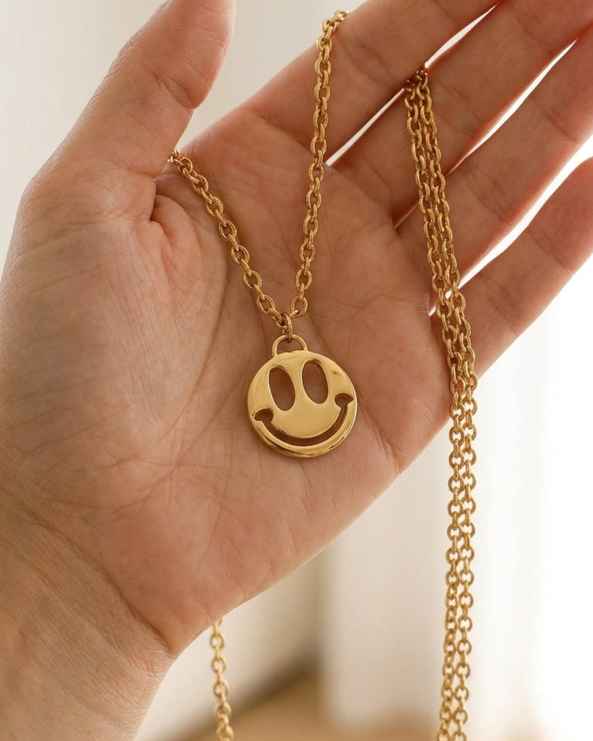 Gold chain necklace featuring a cut-out smile face pendant held in an open hand.