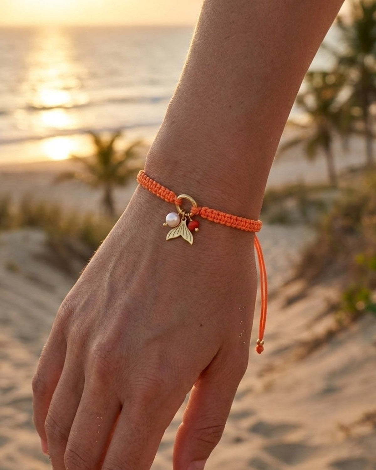 Orange woven cord bracelet with a gold mermaid tail charm and pearl beads on a wrist against a beach backdrop.