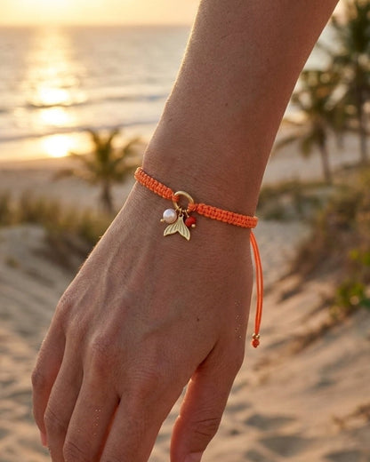 Orange woven cord bracelet with a gold mermaid tail charm and pearl beads on a wrist against a beach backdrop.