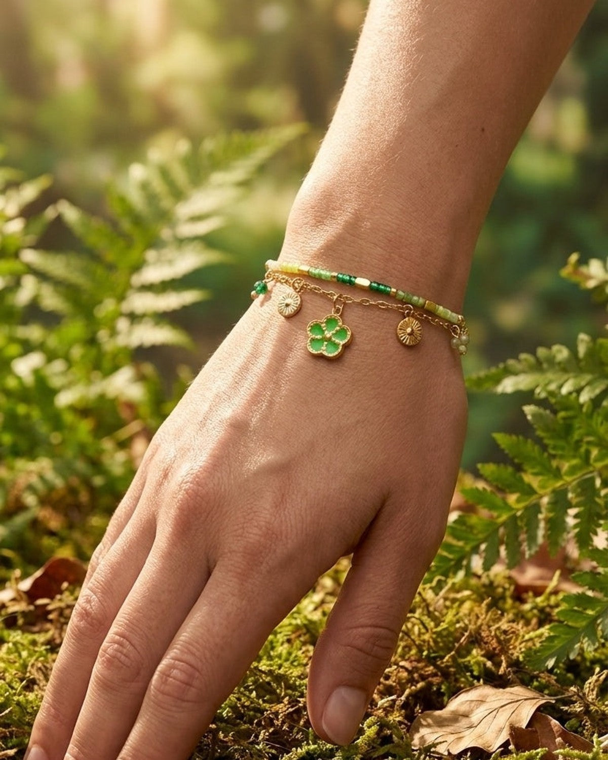 Gold charm bracelet with a green flower charm and sun discs, worn on a wrist over moss.