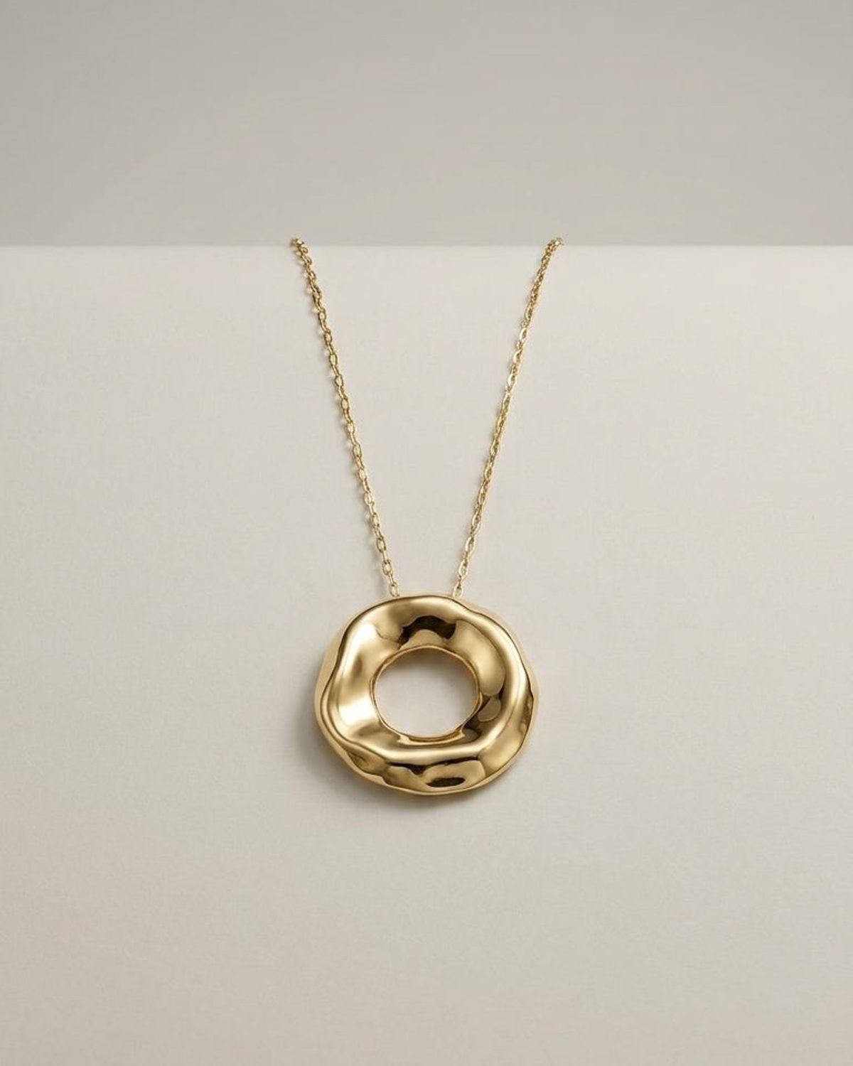 Gold pendant necklace with an uneven, wavy circular ring hanging on a thin chain against a light background.