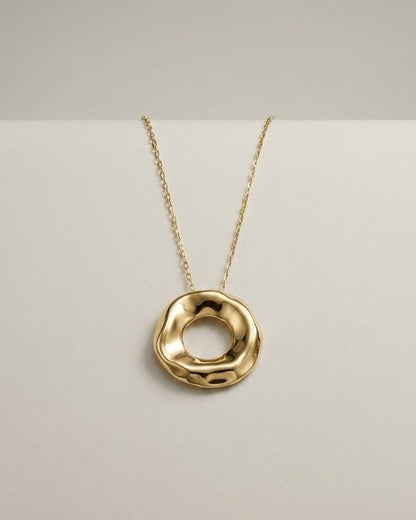 Gold pendant necklace with an uneven, wavy circular ring hanging on a thin chain against a light background.