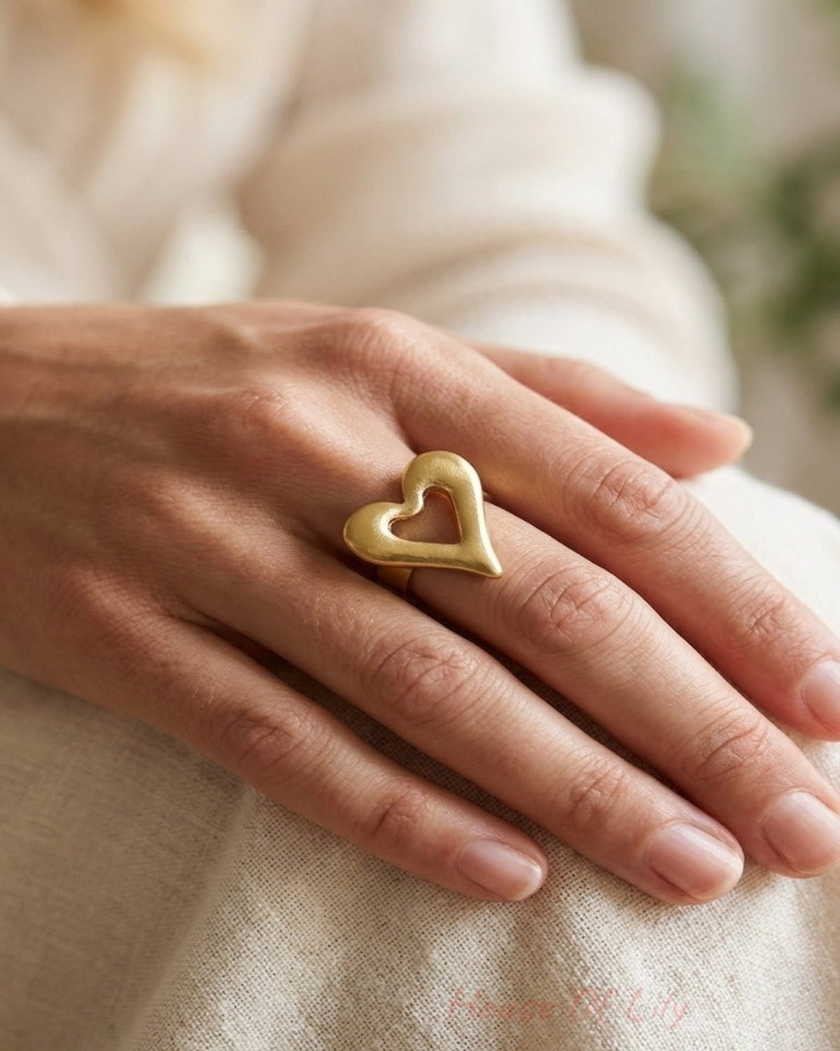 A person wearing a large, brushed gold heart-shaped ring on their finger.