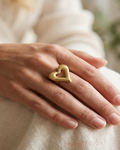 A person wearing a large, brushed gold heart-shaped ring on their finger.