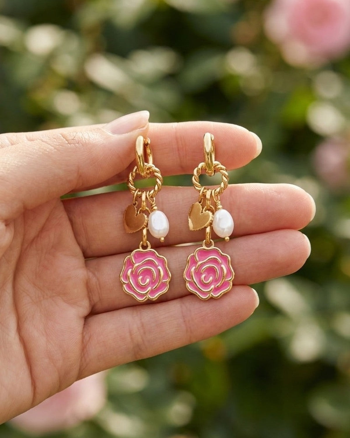 Golden Rose Bloom drop earrings held in hand, featuring gold metal, pink enamel rose, heart, and white pearl charms.