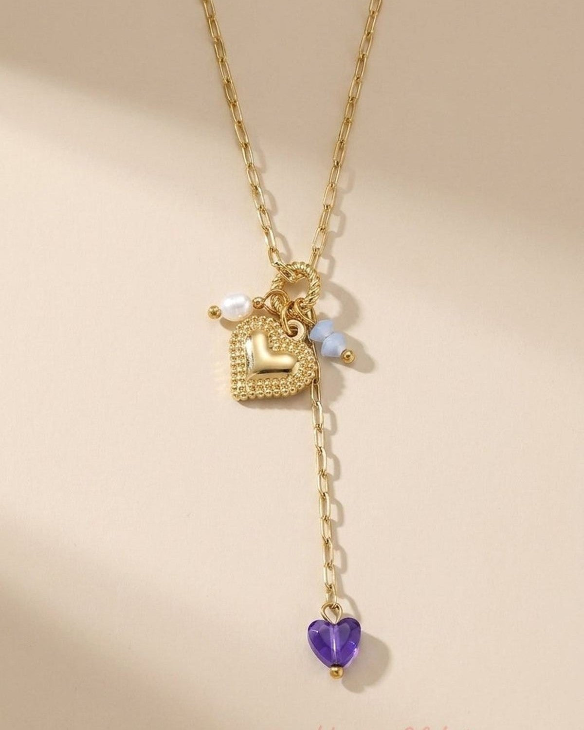 Gold link y-necklace with a textured heart pendant, a white pearl, two blue beads, and a purple heart charm.