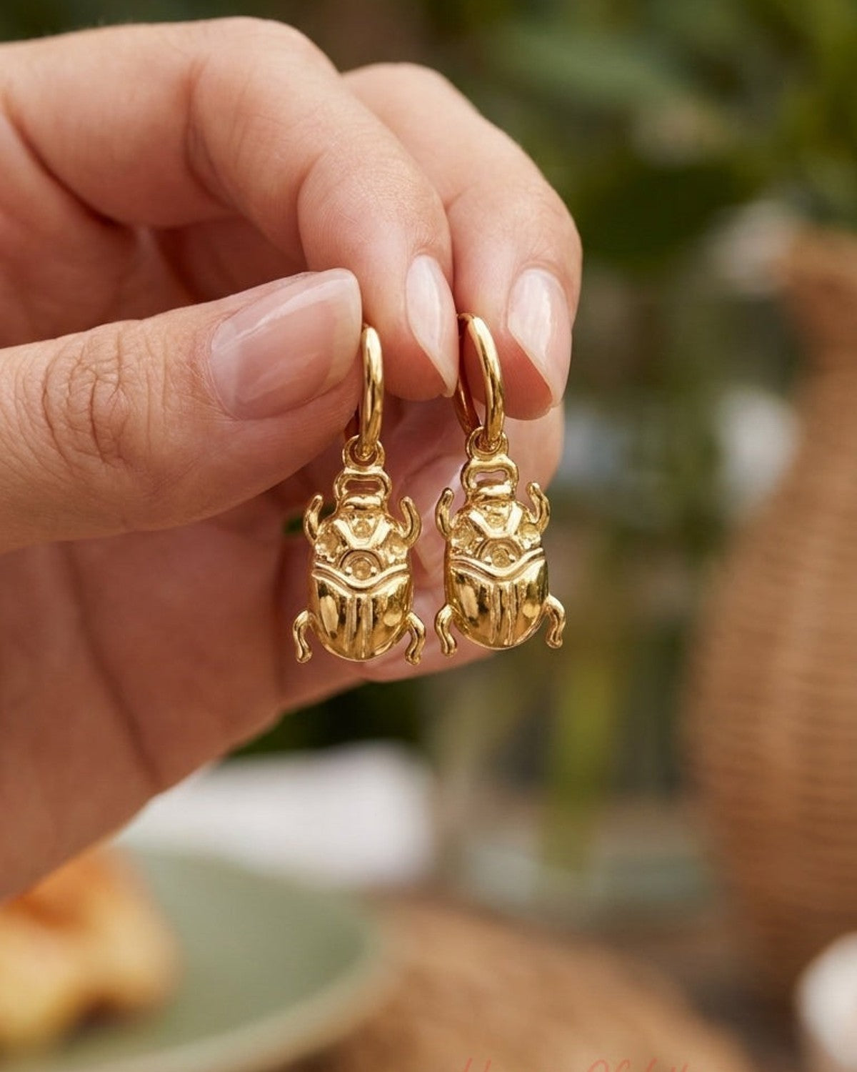 House Of Lily gold drop earrings shaped like polished scarab beetles, held against a blurred background.