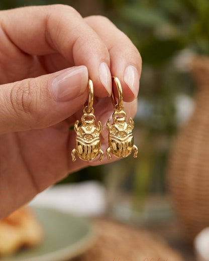House Of Lily gold drop earrings shaped like polished scarab beetles, held against a blurred background.