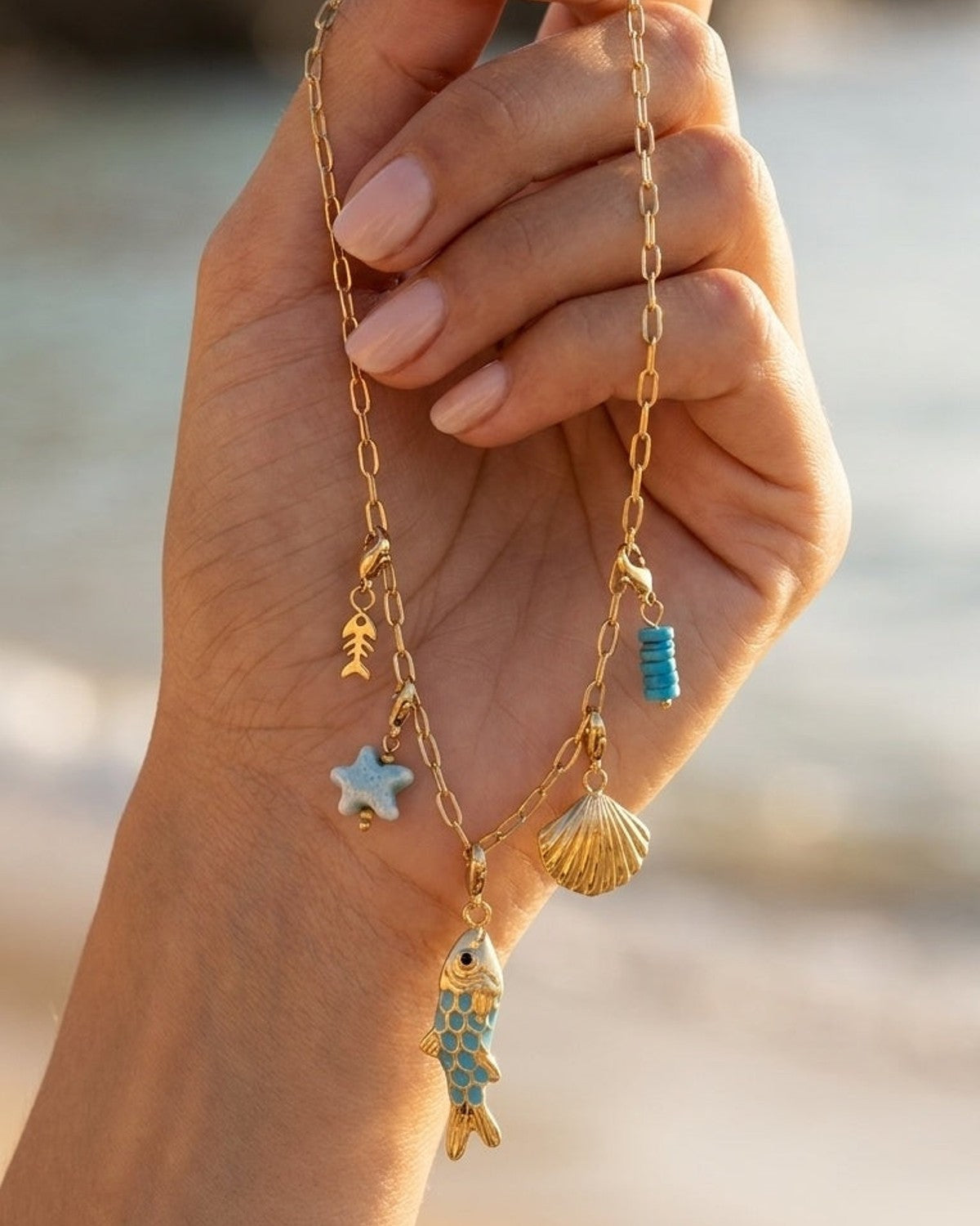 Gold chain bracelet with sea-themed charms: fish, seashell, starfish, and turquoise beads, held by a hand.