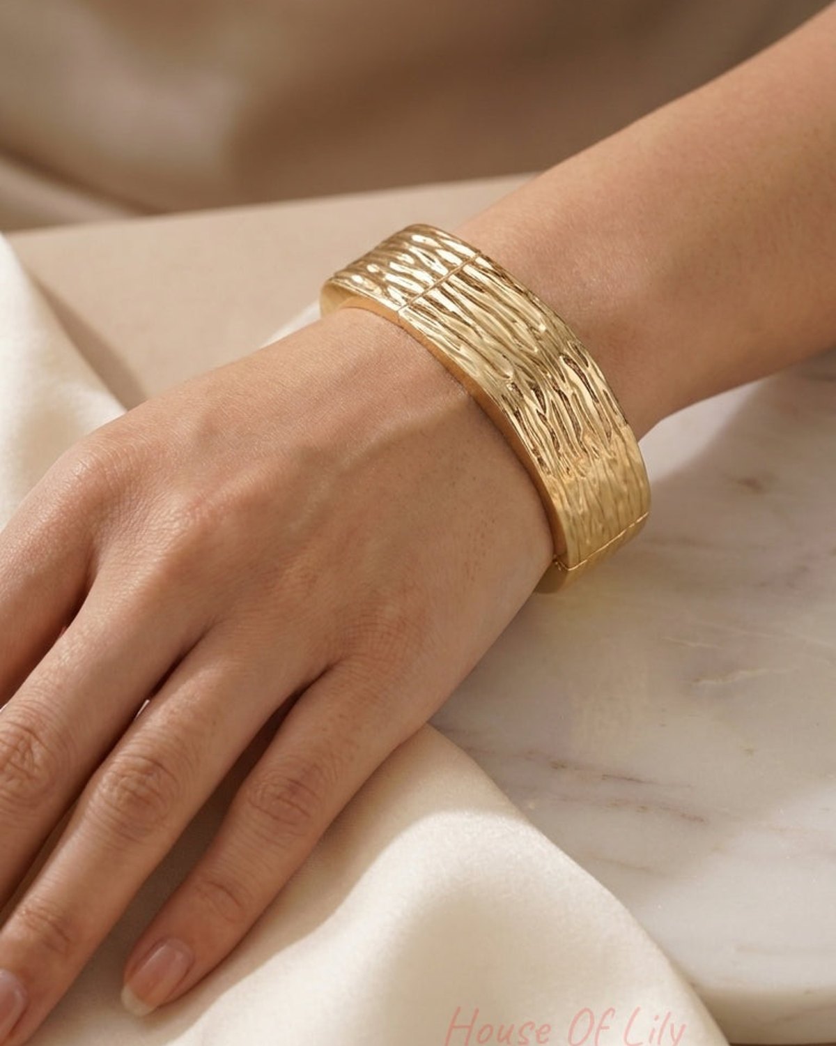 A wide gold hinged bangle with a textured, rippled surface on a woman's wrist.