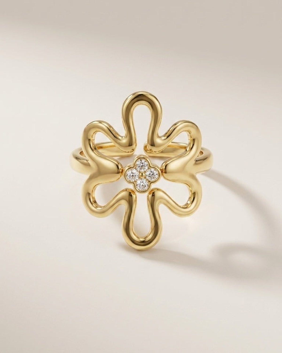 A gold ring shaped like a stylized flower with a small cluster of clear stones at the center, photographed on a clean light beige background.