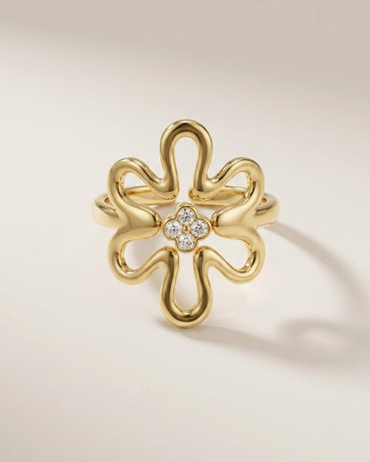 A gold ring shaped like a stylized flower with a small cluster of clear stones at the center, photographed on a clean light beige background.