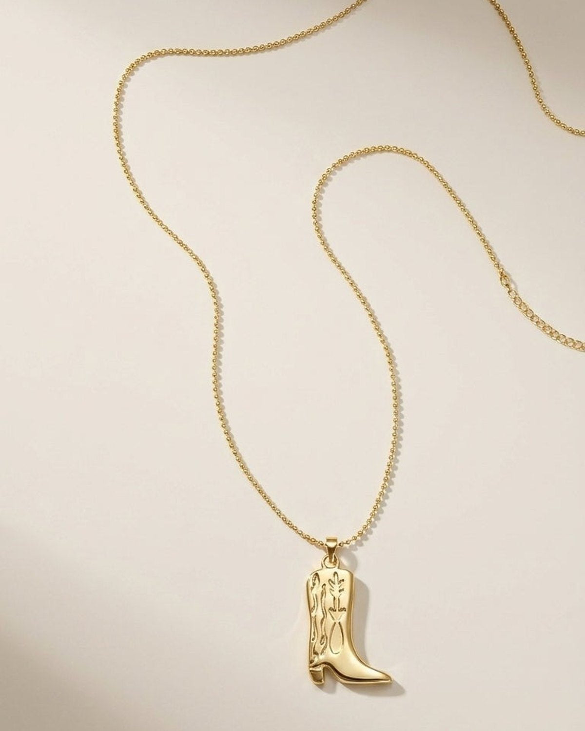 A gold-colored necklace with a cowboy boot pendant lies against a neutral background.