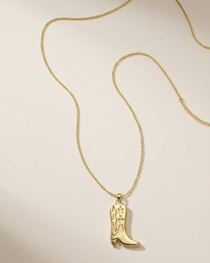 A gold-colored necklace with a cowboy boot pendant lies against a neutral background.