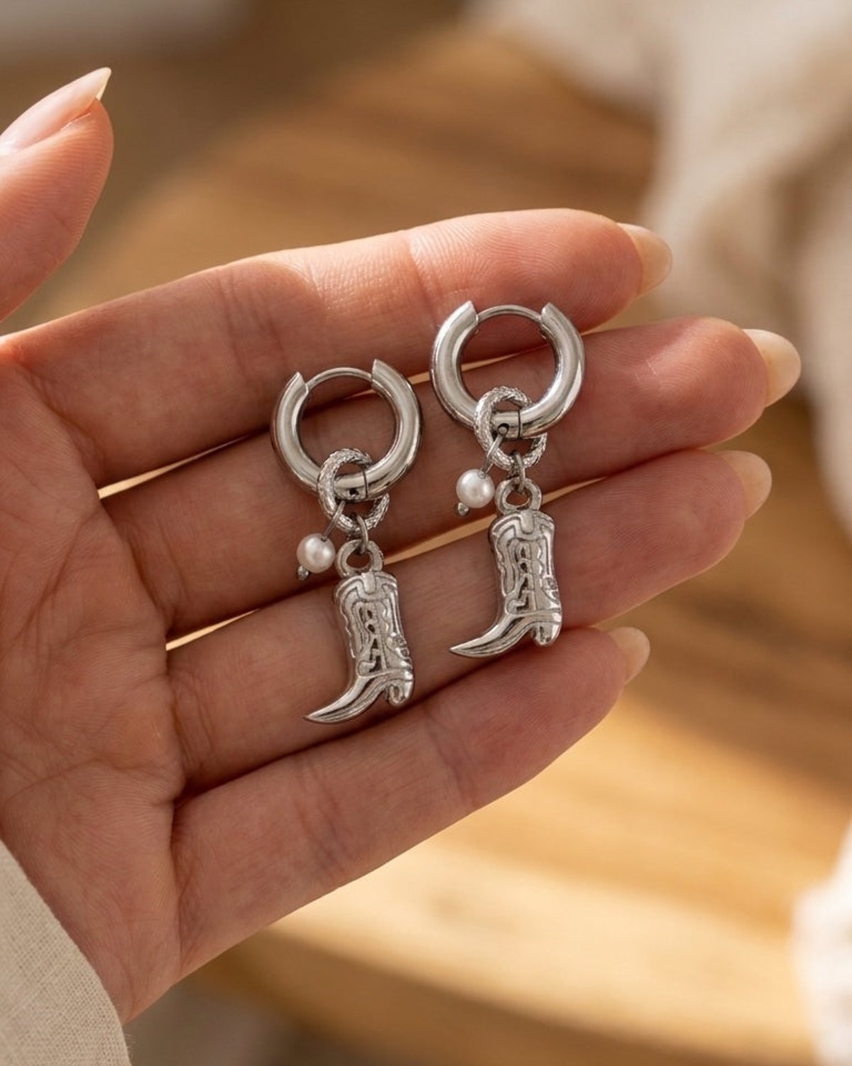 Silver Western cowgirl boot drop earrings with small pearl charms held in an open hand.