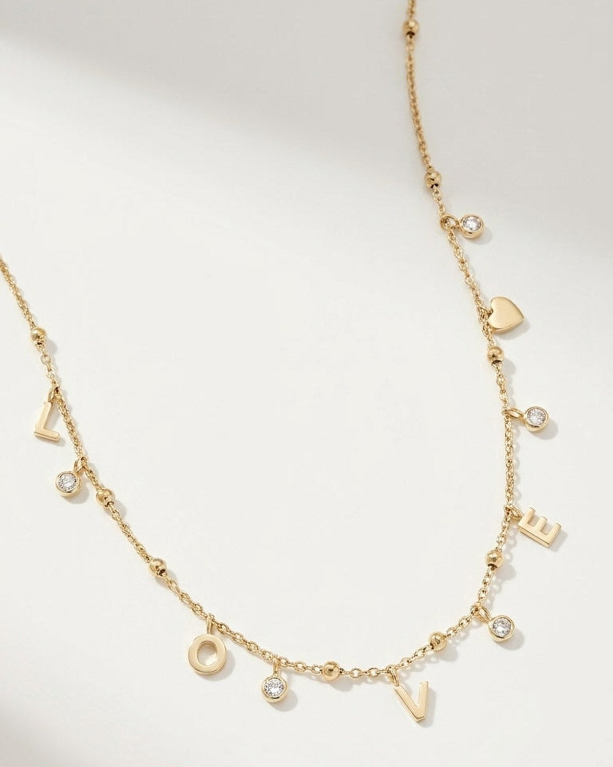 Gold letter pendant necklace spelling "LOVE" with dangling cubic zirconia stones and a small heart charm.