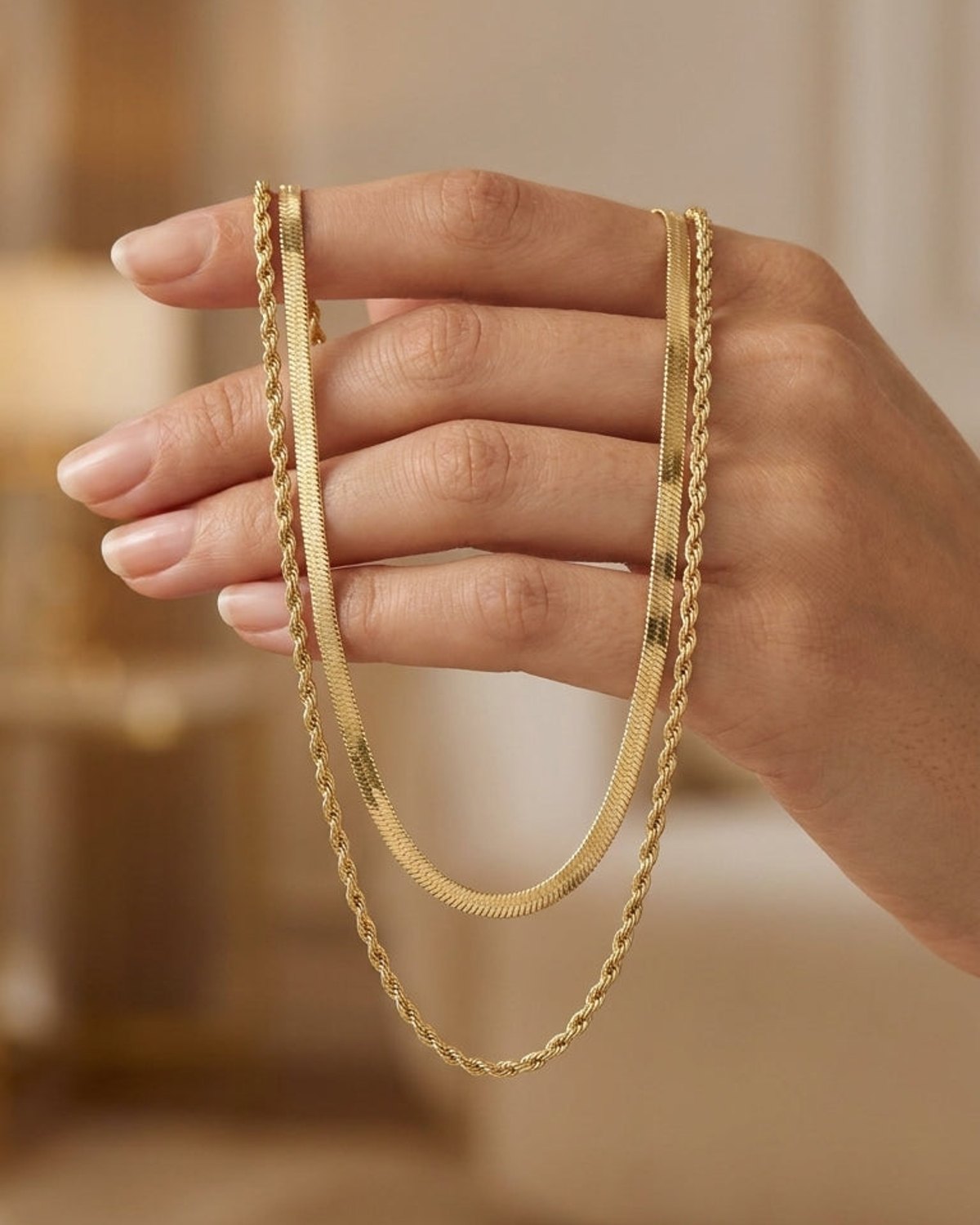 A hand holds a dual gold-toned chain necklace with one flat herringbone chain and one textured rope chain.