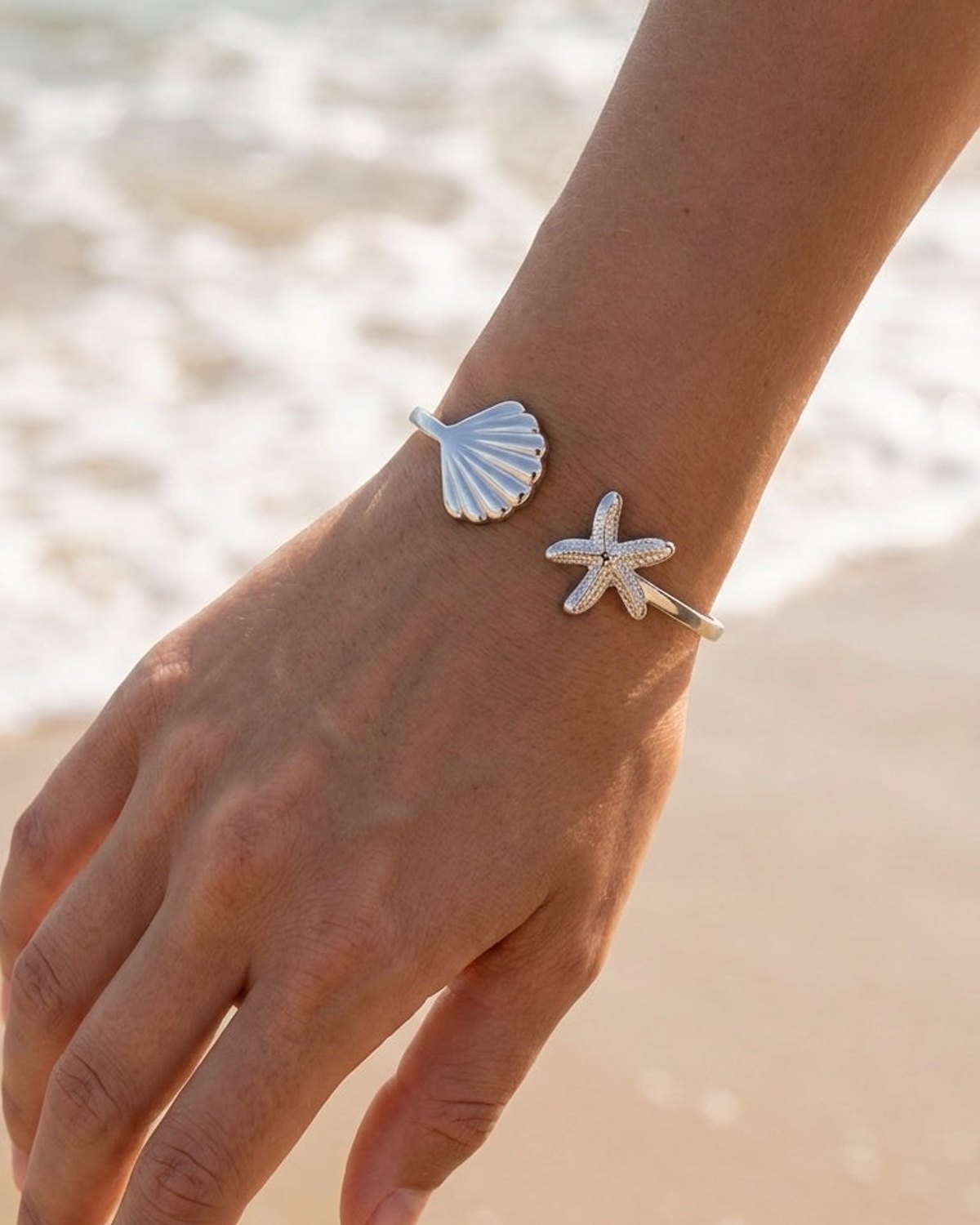 A silver cuff bracelet with a shell and a pavé-set crystal starfish on a woman's wrist at a beach.