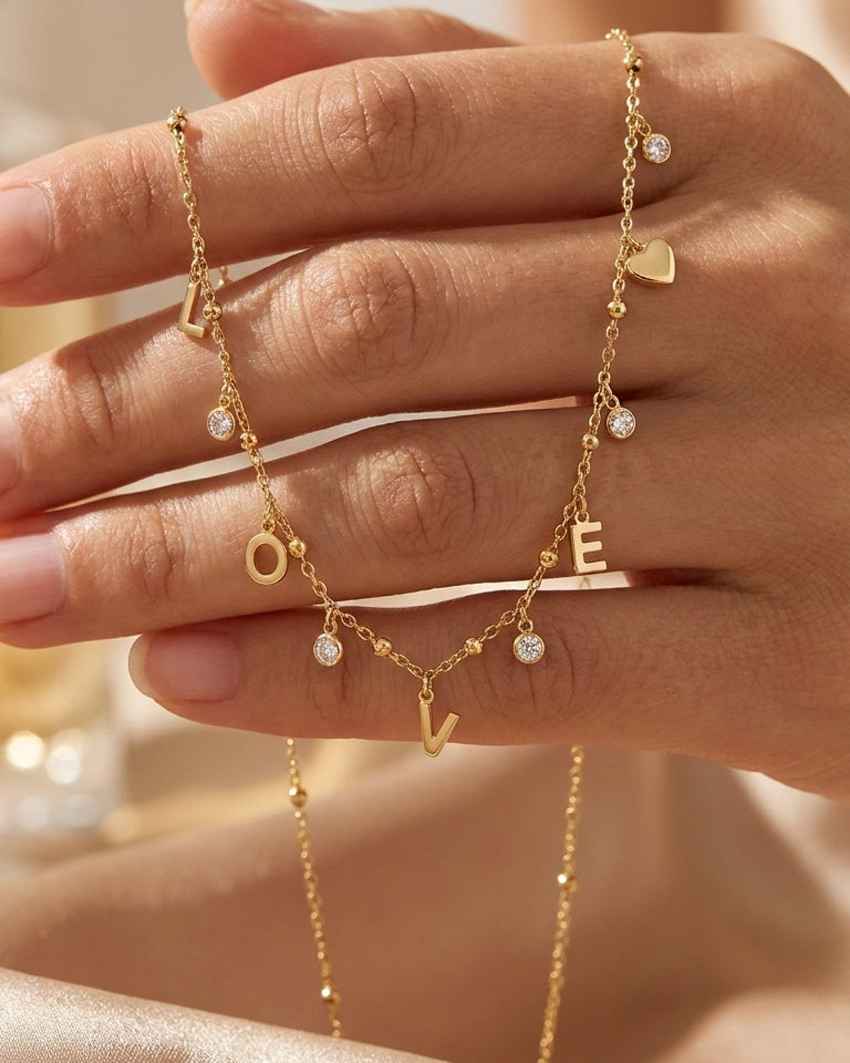 Gold Letter Pendant Necklace draped across fingers, featuring dangling "LOVE" letter charms, rhinestones, and a small heart.
