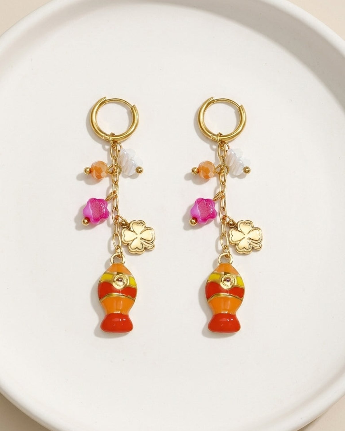 Gold hoop dangle earrings with enamel orange fish charms, flower beads, and clover accents on a white surface.