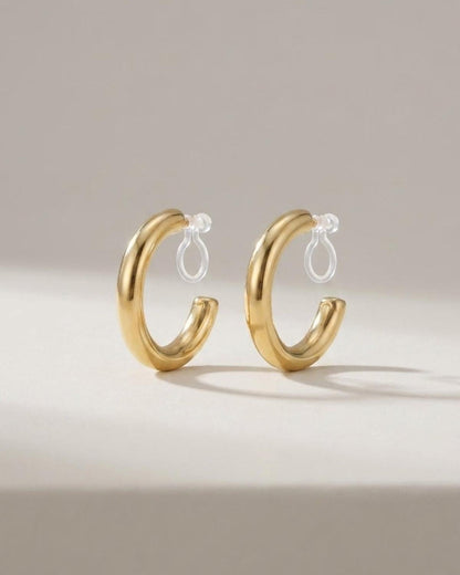 Thick gold hoop earrings with clear clip-on backs, displayed standing upright on a plain surface.