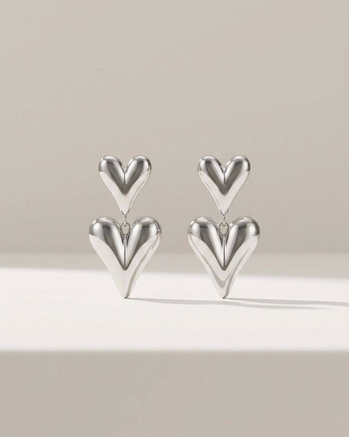 Two silver drop earrings featuring puffed, stacked heart charms on a neutral background.