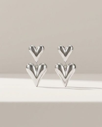 Two silver drop earrings featuring puffed, stacked heart charms on a neutral background.