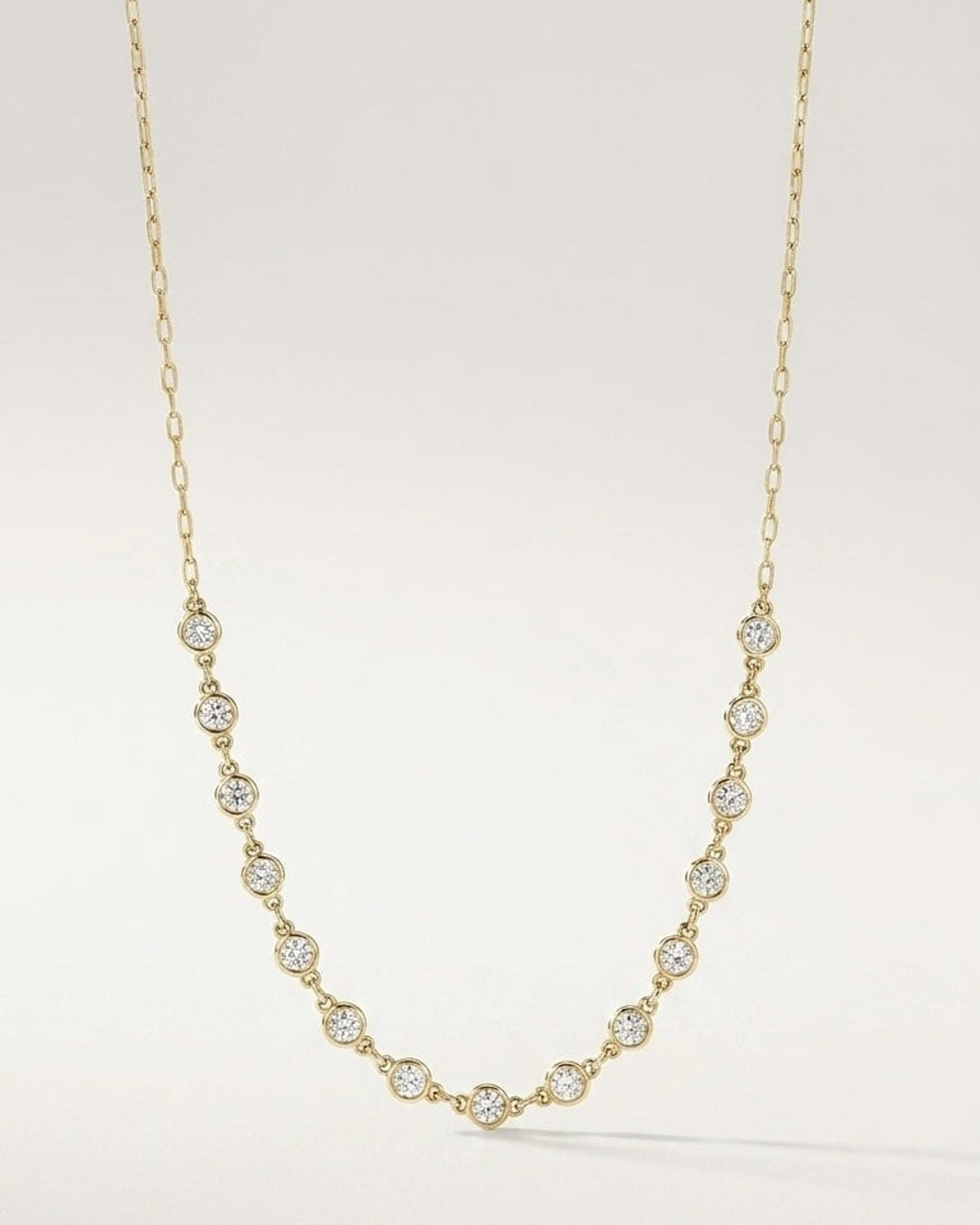 Gold chain necklace featuring evenly spaced round bezel-set stones along the lower drop against a plain background.