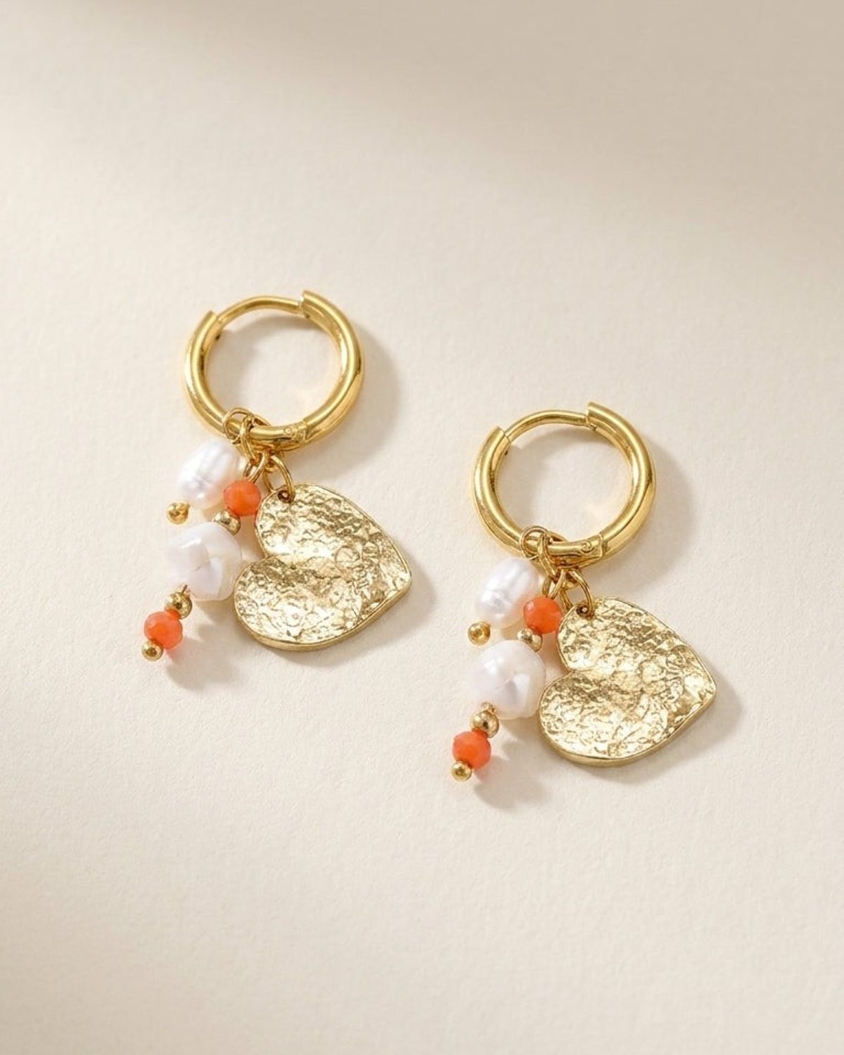 Gold hoop earrings with textured heart charms, white pearls, and orange beads on a neutral background.