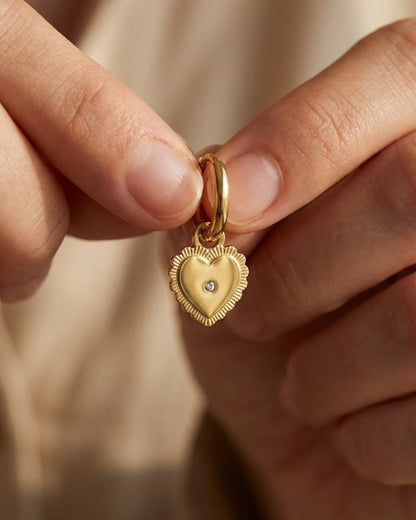 Gold-plated drop earring featuring a puffy heart charm with a scalloped edge and a central crystal, held in fingers.