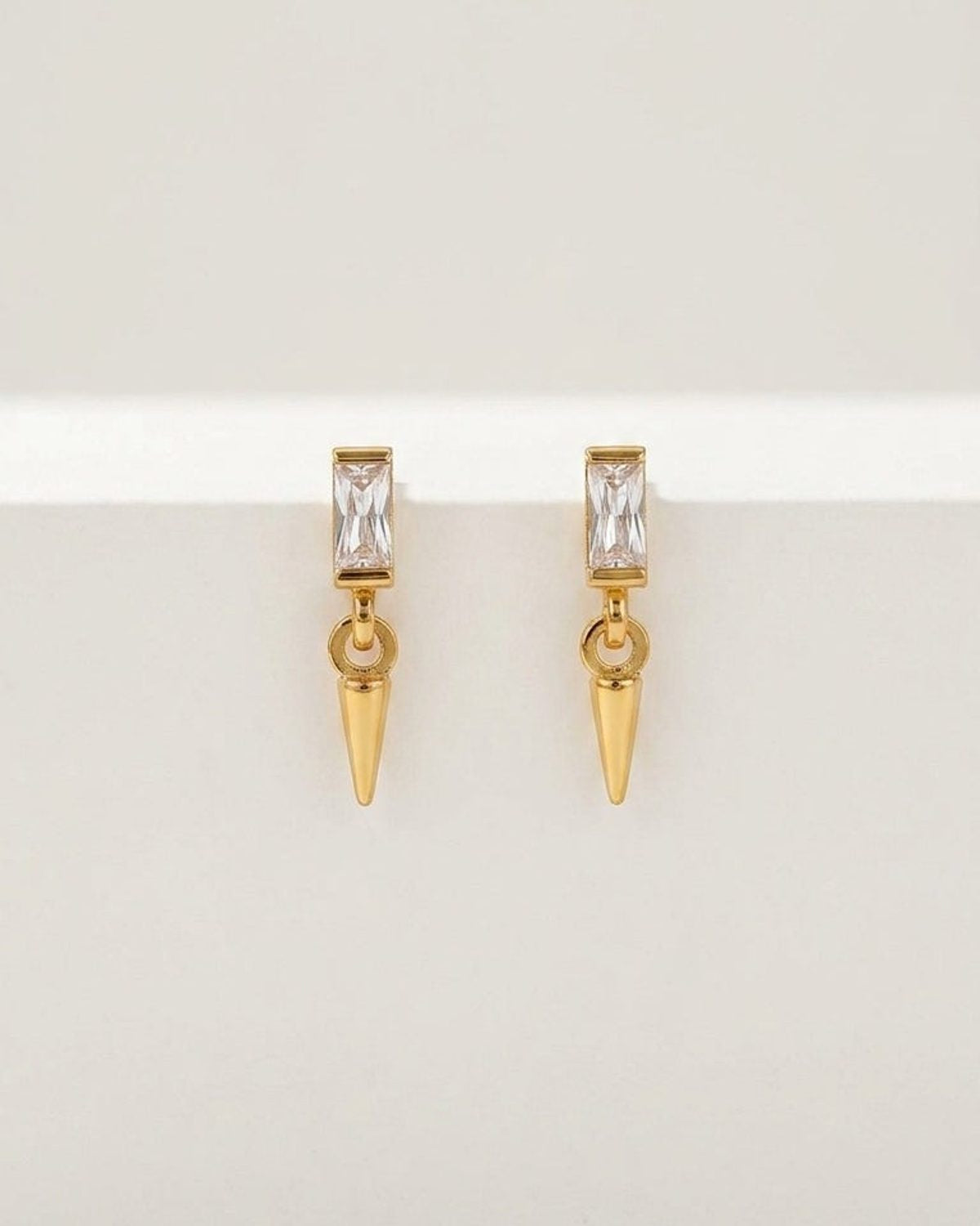 Gold-toned drop earrings featuring a rectangular crystal stud and a dangling small cone pendant on a white surface.