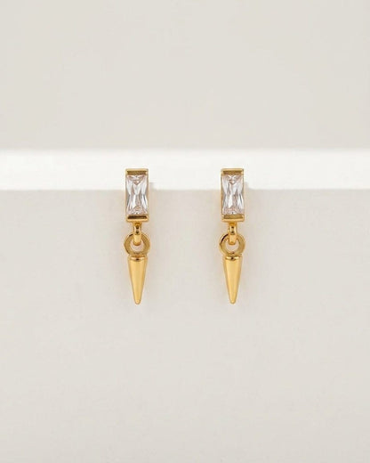 Gold-toned drop earrings featuring a rectangular crystal stud and a dangling small cone pendant on a white surface.