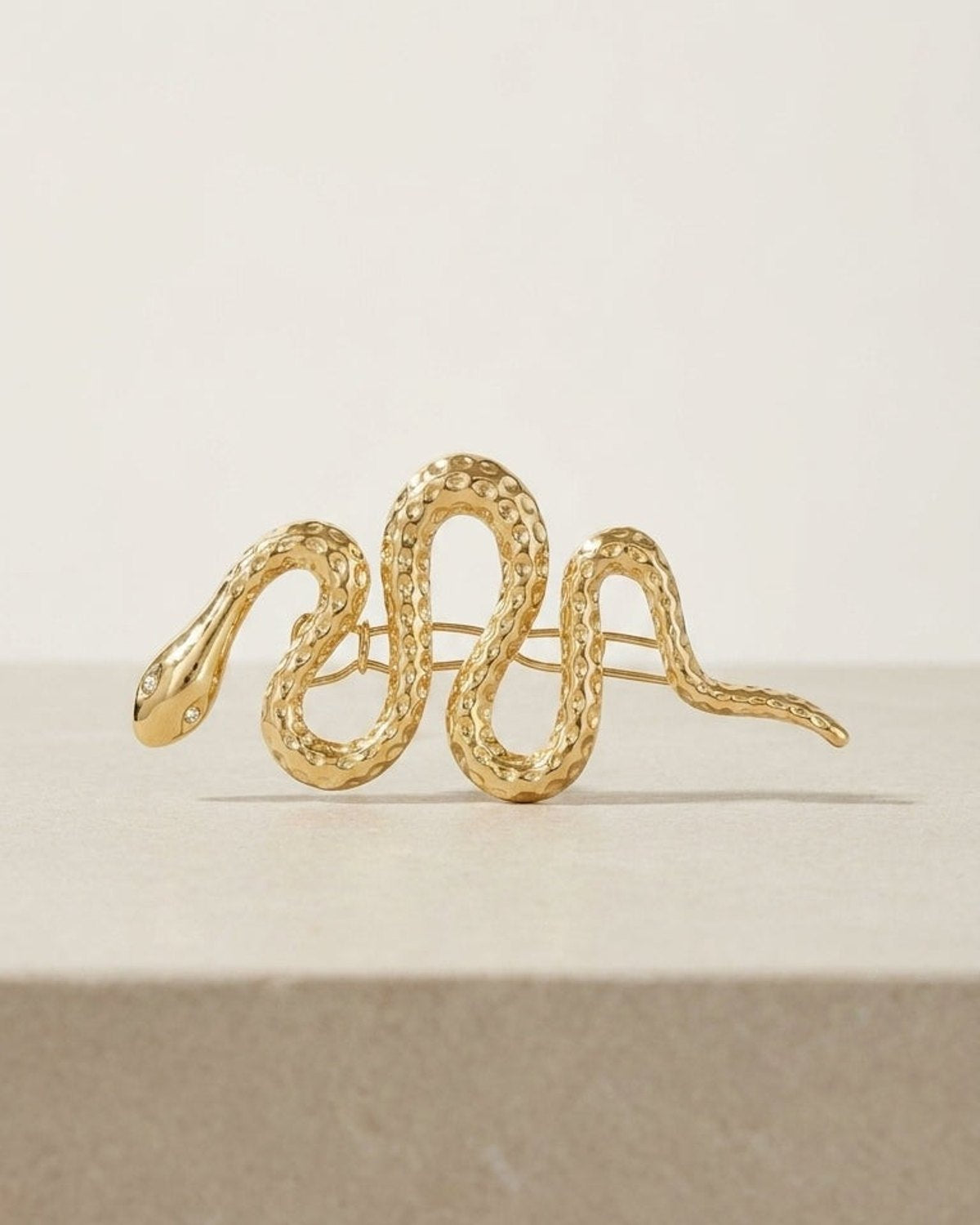 Gold hair clip in a coiled, textured snake shape with crystal eyes, shown on a neutral surface.