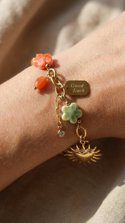 Gold chain charm bracelet with coral-colored stone, green flower, "Good Luck" tag, crystal accent, and sun heart charm.