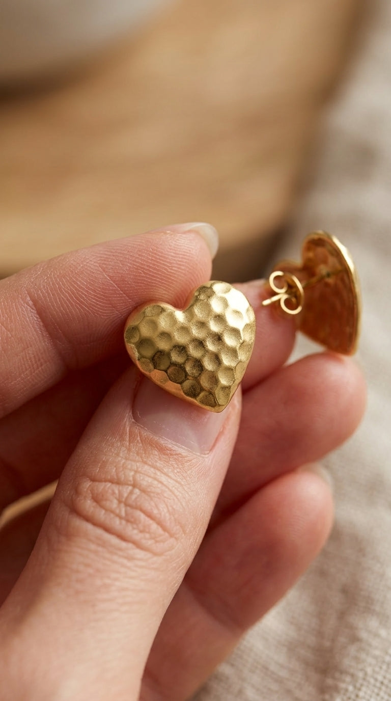 Gold heart-shaped stud earrings with a textured, hammered finish held between fingers.