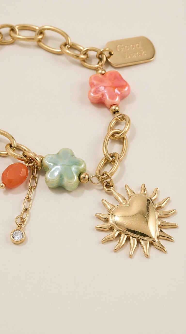 Gold-tone chain charm bracelet featuring a heart-sun pendant, flower charms, Good Luck tag, and a tiny crystal accent.