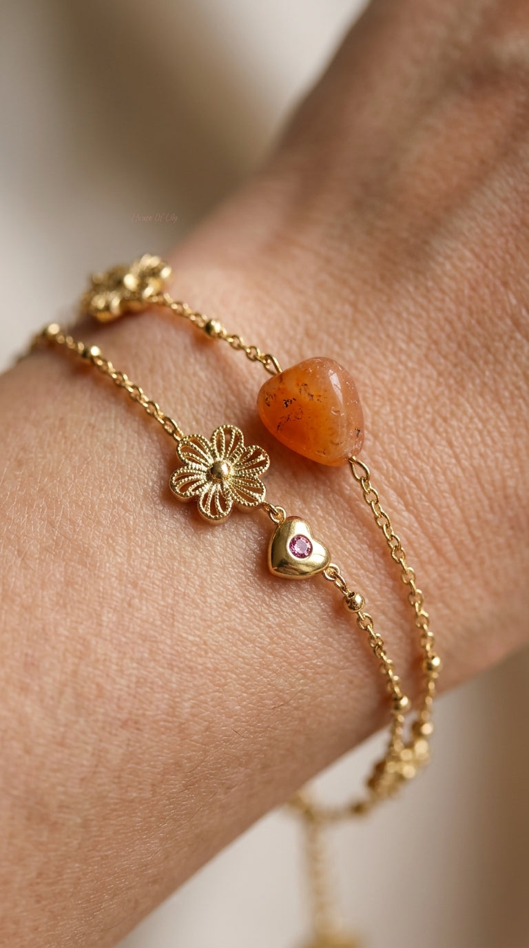 Gold-tone layered charm bracelets featuring a carnelian stone, flower charm, and a heart with a pink gemstone.