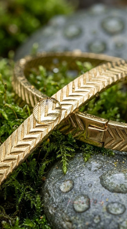 Gold-toned herringbone bracelet with a sleek hinge clasp, resting on damp moss and smooth, wet river stones.