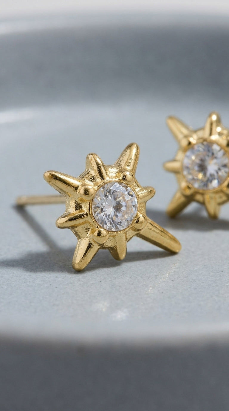 Gold starburst stud earrings featuring a central round-cut clear crystal, set in a textured, polished gold finish.