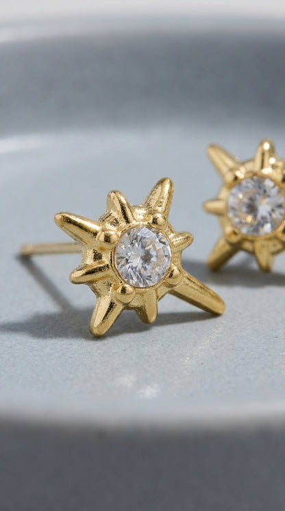 Gold starburst stud earrings featuring a central round-cut clear crystal, set in a textured, polished gold finish.