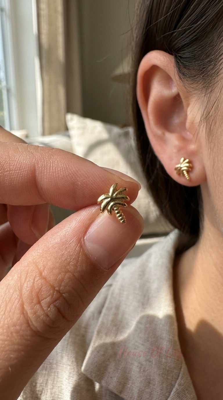 Gold-tone palm tree stud earrings with polished finish, shown being held and worn in an earlobe.