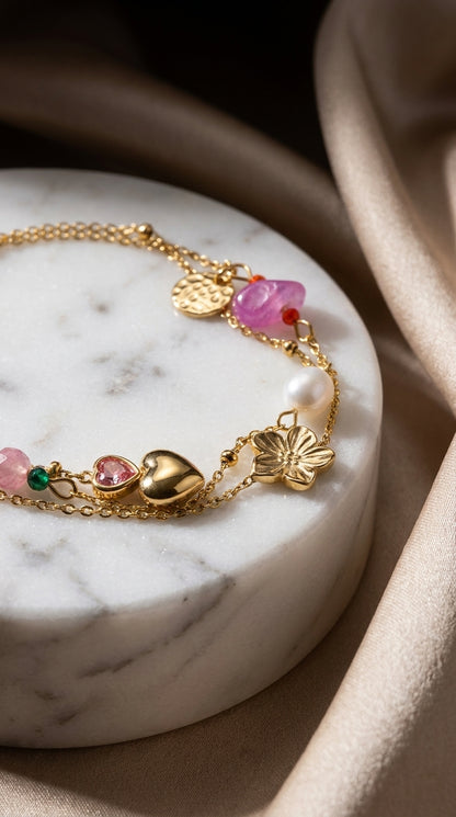 Gold-tone charm bracelet with heart, flower, and coin charms featuring colorful gemstones and a white pearl.